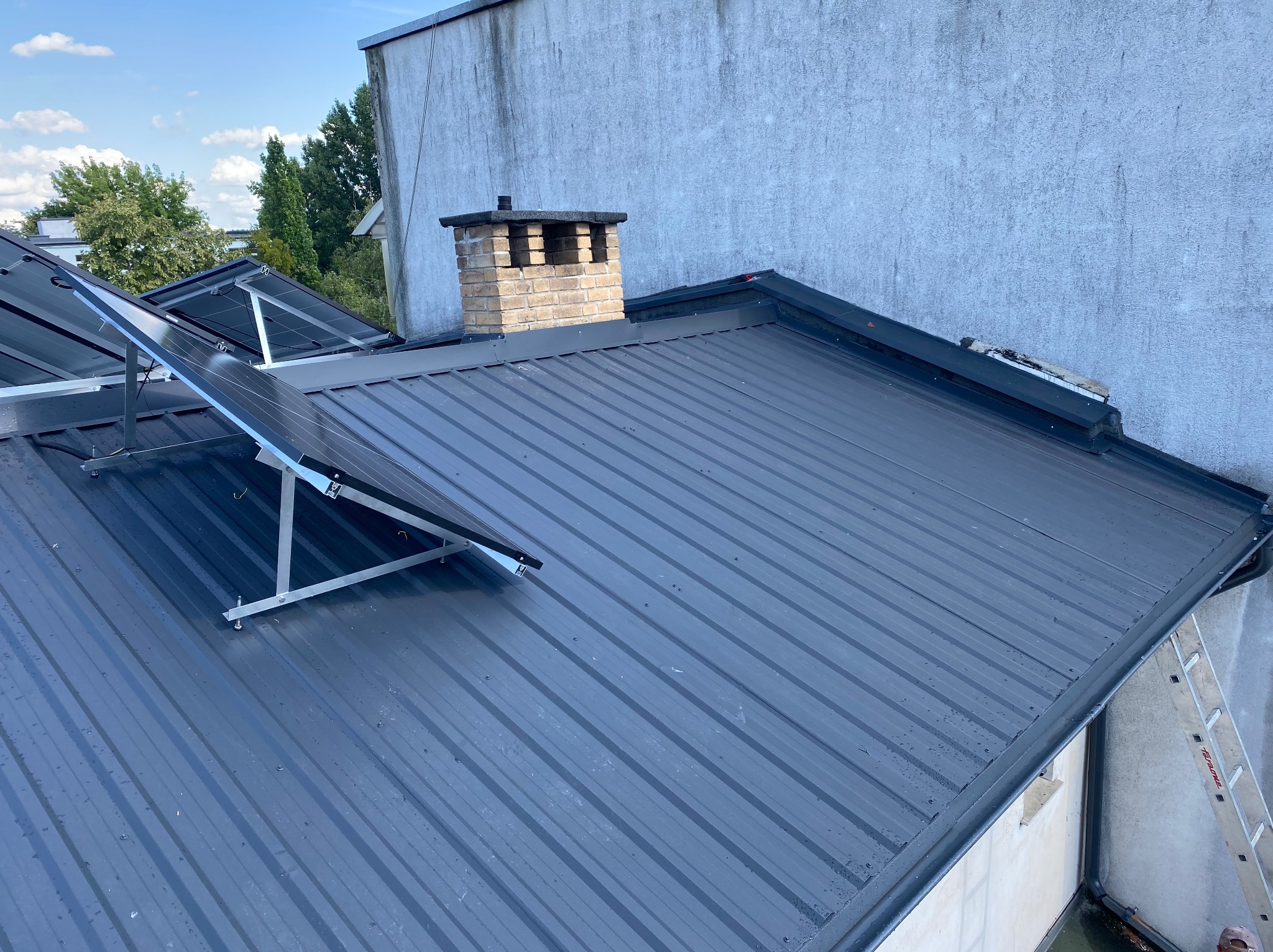 Nowoczesny, grafitowy dach z blachy trapezowej z zamontowanymi panelami fotowoltaicznymi i ceglanym kominem na tle jasnej ściany budynku.