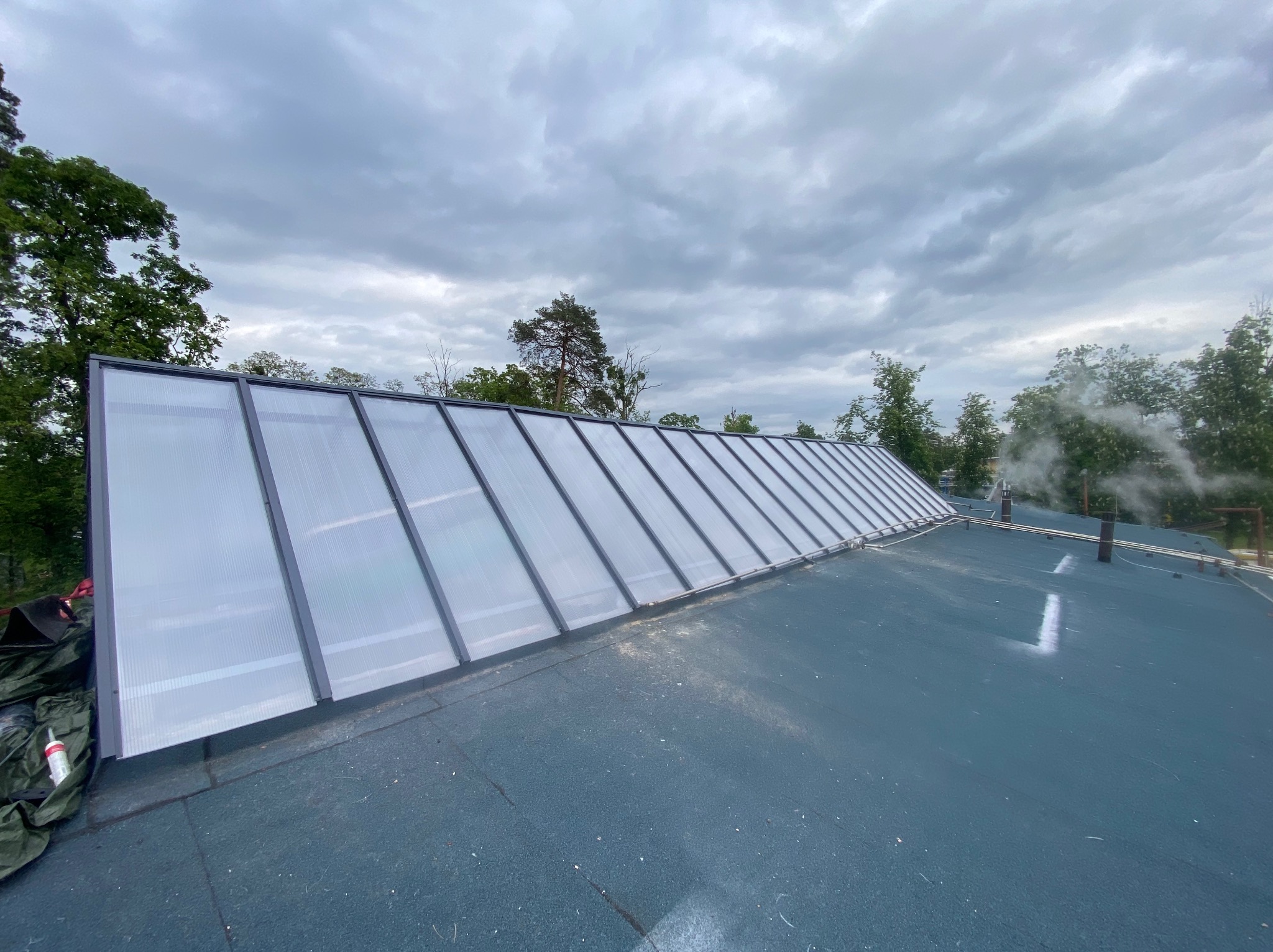 Dach pokryty papą z zamontowanym świetlikiem dachowym z szarymi profilami. W tle zieleń drzew i pochmurne niebo. Widoczny dym z komina.