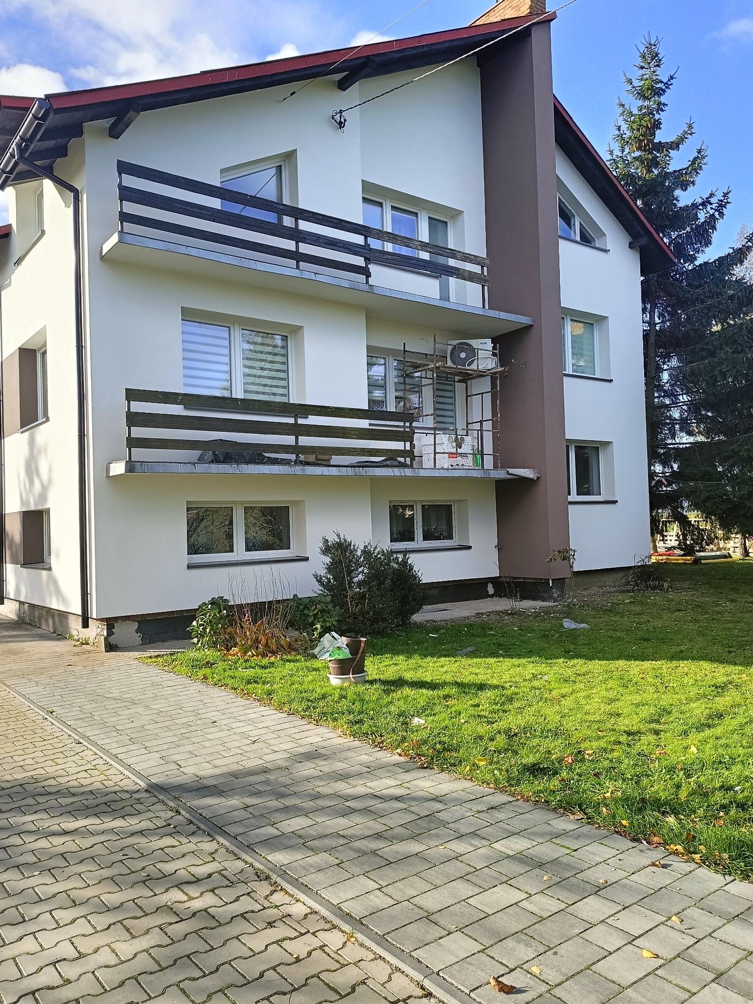 Dwupiętrowy dom po renowacji elewacji, z balkonami i brązowym akcentem kolumnowym. Widoczne rusztowania na jednym z balkonów. Zieleń trawnika i kostka brukowa.