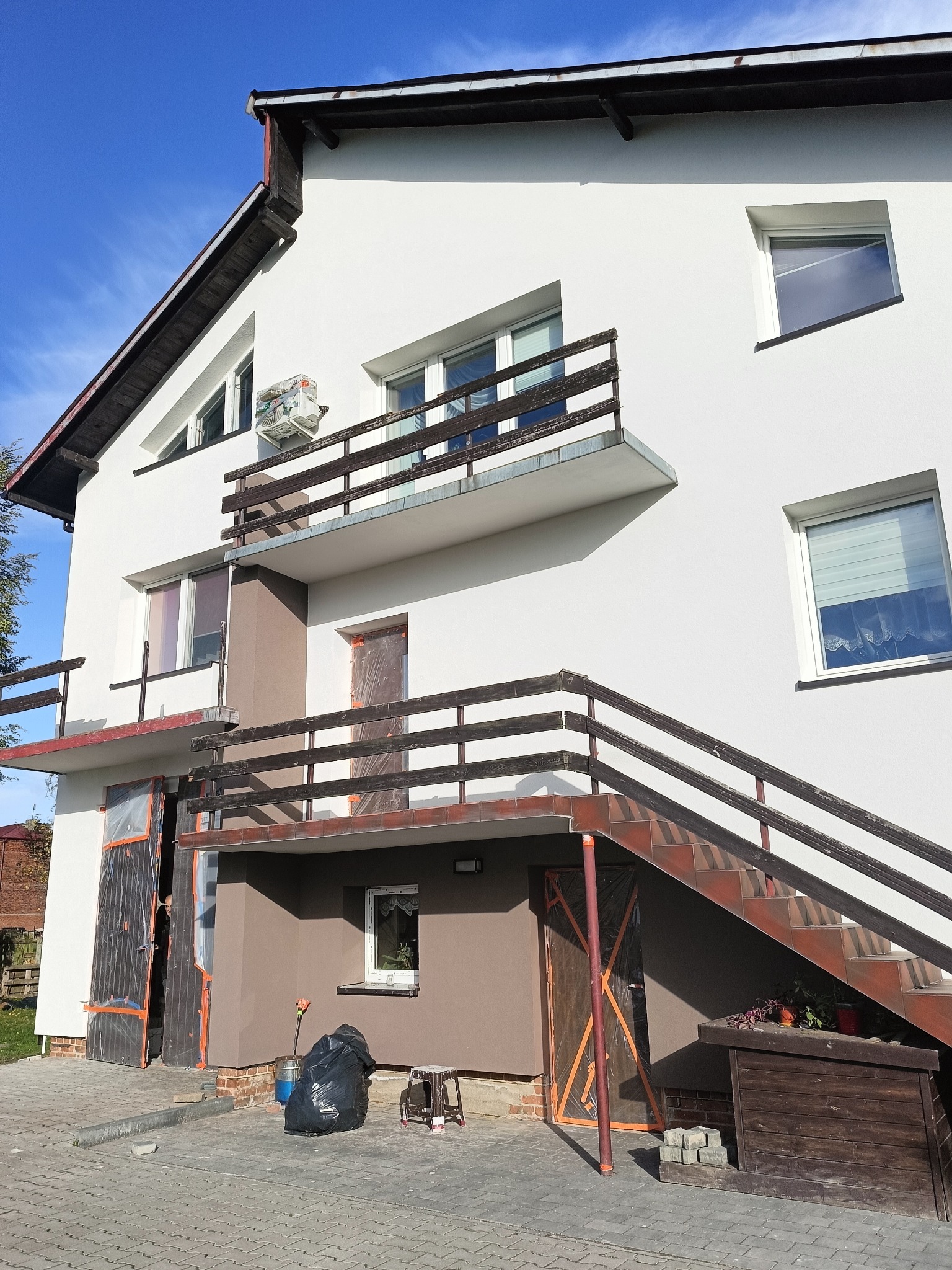 Elewacja domu po malowaniu w Piotrków Trybunalski. Jasna fasada z brązowymi akcentami, widoczne zabezpieczone okna i drzwi, balkon z drewnianą balustradą.