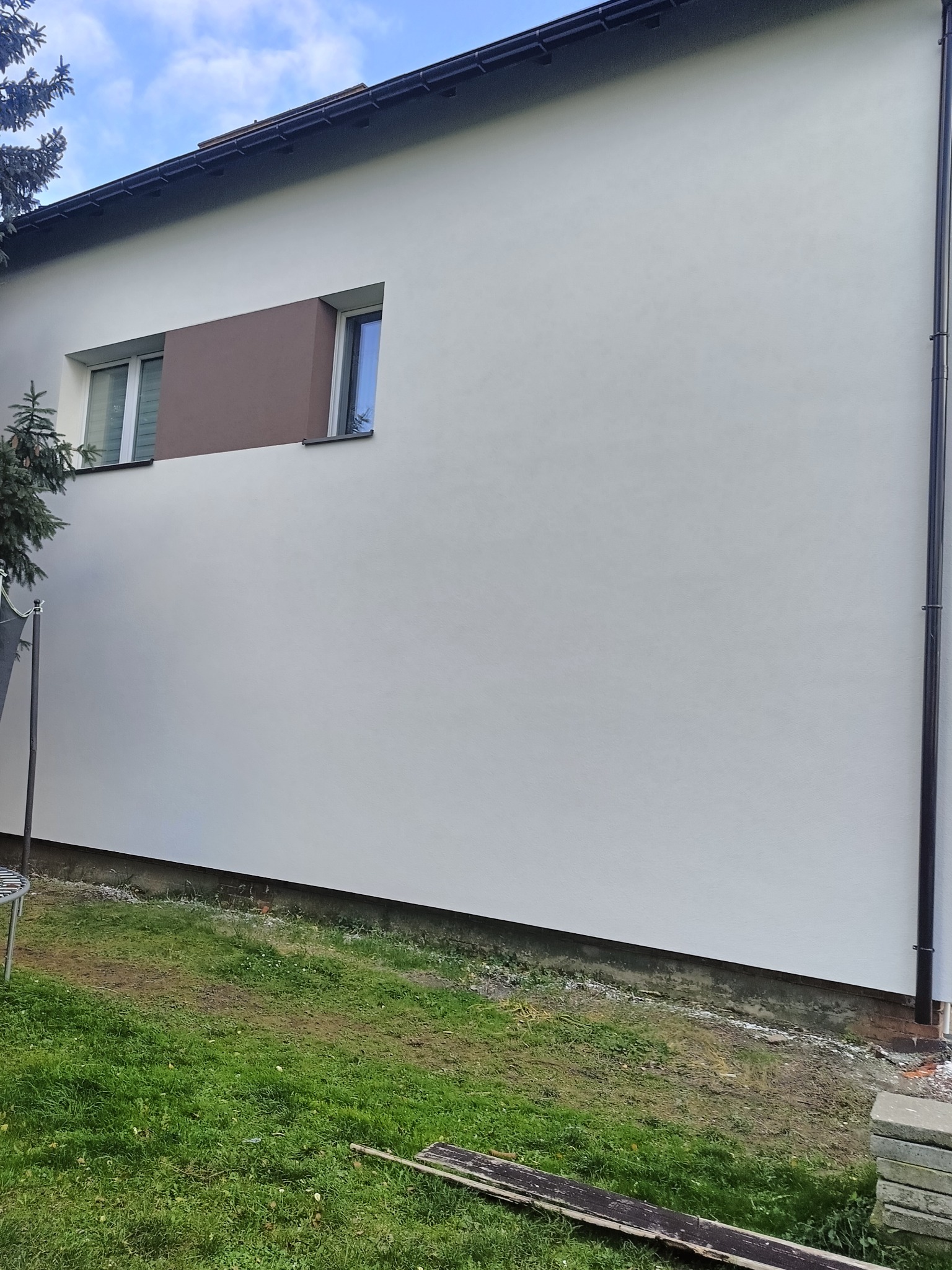 Elewacja domu po malowaniu na biało z brązowym akcentem wokół okna. Widoczny fragment ogrodu i rynna. Minimalistyczny design.