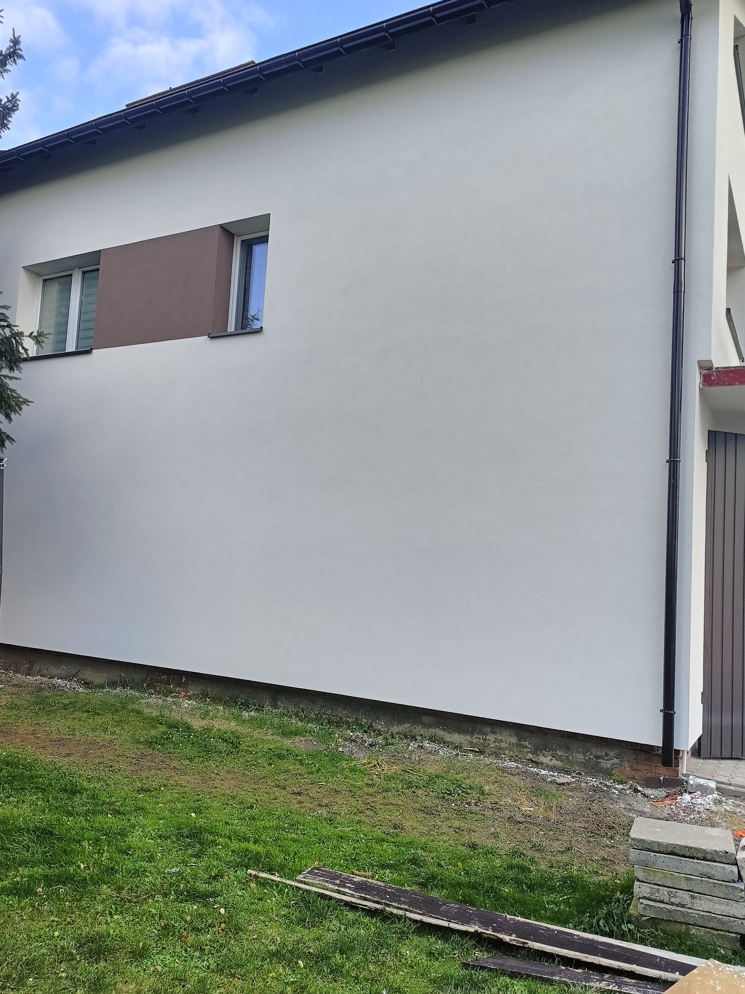 Biała fasada budynku po renowacji z brązowym akcentem wokół okna. Widoczny fragment dachu, rynna i trawnik. Świeżo pomalowana ściana.