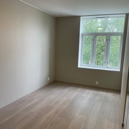 Jasne, puste wnętrze pokoju z drewnianą podłogą i dużym oknem z widokiem na zieleń. Ściany w odcieniach beżu, minimalistyczne wykończenie, nowoczesne oświetlenie sufitowe.