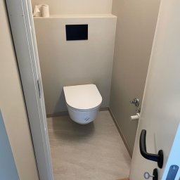 Nowoczesna, minimalistyczna łazienka z podwieszaną toaletą i beżowymi płytkami. Widok zza uchylonych drzwi, akcent na czarny panel spłuczki.