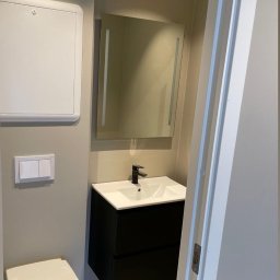 Wykończona łazienka w neutralnych barwach: podwieszana toaleta, czarna szafka z umywalką, lustro z oświetleniem LED i białe drzwi. Widok po remoncie.