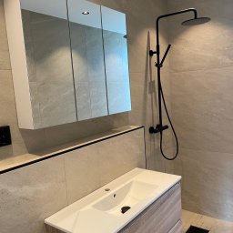 Nowoczesna łazienka z beżowymi płytkami, czarną armaturą i szafką z lustrem. Umywalka z drewnopodobną szafką. Minimalistyczny design, oświetlenie punktowe.