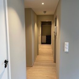 Proff Tools - Wykończony korytarz w odcieniach szarości i beżu z drewnianą podłogą. Widoczne drzwi, włącznik światła i gniazdko elektryczne. W głębi szafa z czarnymi półkami.