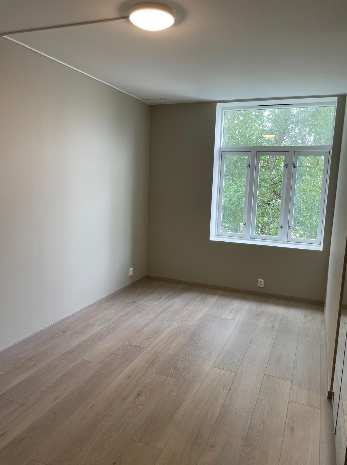 Jasne, puste wnętrze pokoju z drewnianą podłogą i dużym oknem z widokiem na zieleń. Ściany w odcieniach beżu, minimalistyczne wykończenie, nowoczesne oświetlenie sufitowe.