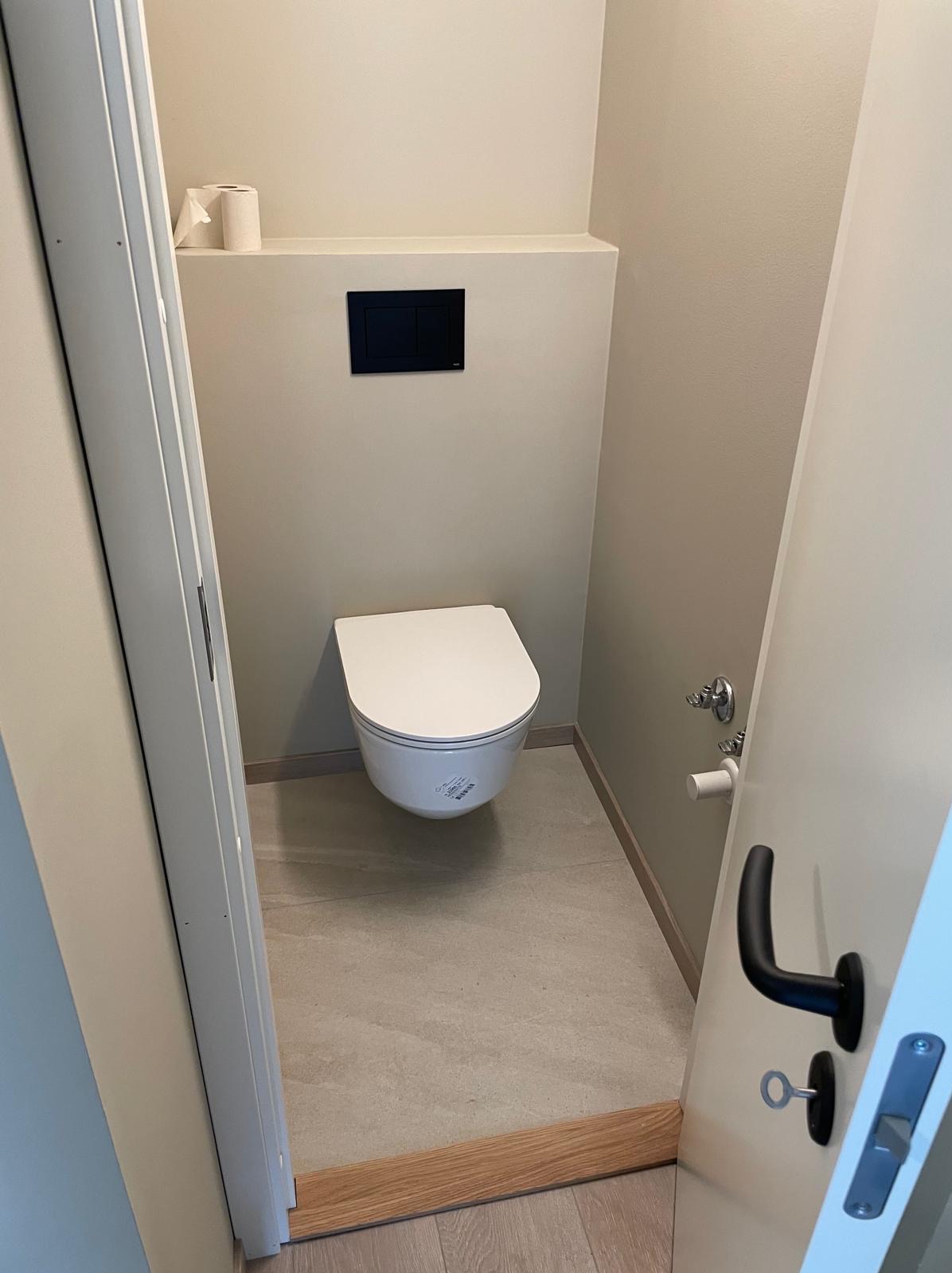 Nowoczesna, minimalistyczna łazienka z podwieszaną toaletą i beżowymi płytkami. Widok zza uchylonych drzwi, akcent na czarny panel spłuczki.