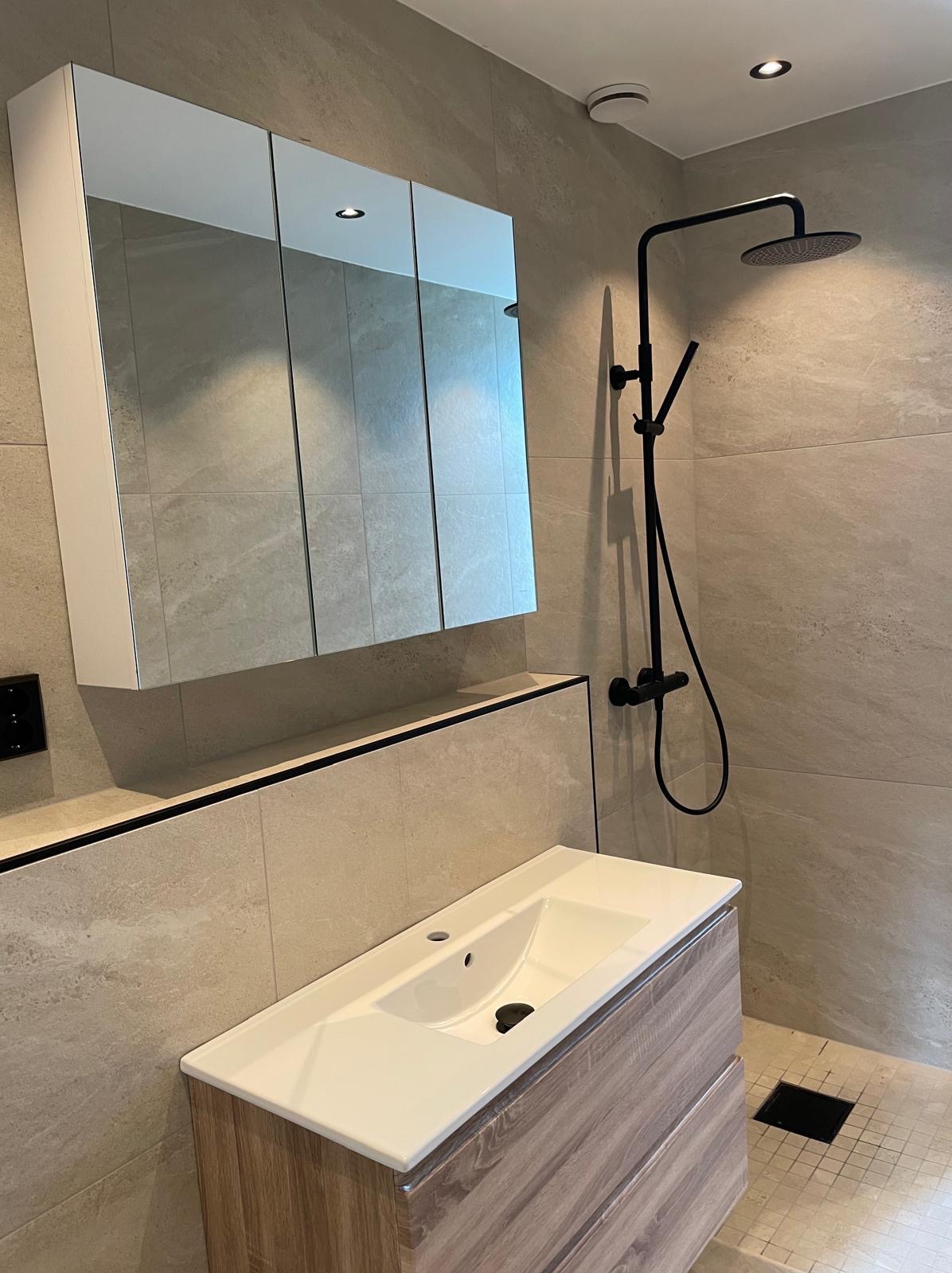 Nowoczesna łazienka z beżowymi płytkami, czarną armaturą i szafką z lustrem. Umywalka z drewnopodobną szafką. Minimalistyczny design, oświetlenie punktowe.