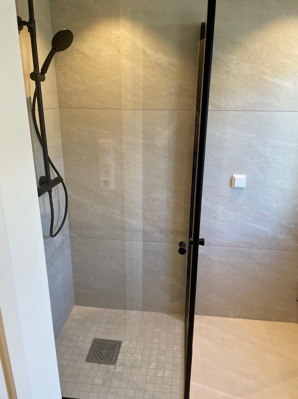 Nowoczesna kabina prysznicowa z czarną armaturą i odpływem liniowym w posadzce, wykończona płytkami w odcieniach beżu i szarości. Minimalistyczny design.