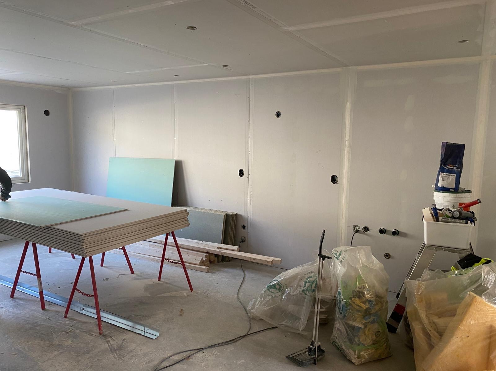 Wnętrze w trakcie montażu płyt kartonowo-gipsowych na ścianach i suficie. Widoczne narzędzia, materiały budowlane i stelaż pod płyty. Otwory pod instalacje elektryczne.