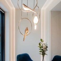 BAMARI Mariola Fatyga - Nowoczesna lampa wisząca z motywem ptaków nad okrągłym stołem i welurowymi krzesłami w jadalni. Minimalistyczny design z akcentami złota i subtelnym oświetleniem.