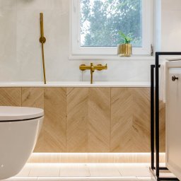 BAMARI Mariola Fatyga - Jasna łazienka z drewnianą obudową wanny ułożoną w jodełkę, złotą armaturą i rośliną w złotej doniczce na parapecie. Widoczna toaleta i szafka łazienkowa.