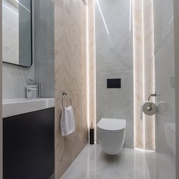 BAMARI Mariola Fatyga - Nowoczesna łazienka z podświetlanymi panelami, umywalką z czarną szafką i wiszącą toaletą. Jasne płytki imitujące drewno i marmur. Minimalistyczny design.