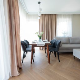 BAMARI Mariola Fatyga - Jasne wnętrze: stół z szarymi krzesłami, sofa, parkiet w jodełkę, delikatne zasłony i nowoczesna lampa. Minimalistyczny design z akcentami beżu i szarości.