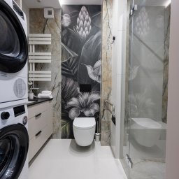 BAMARI Mariola Fatyga - Nowoczesna łazienka z motywem florystycznym na ścianie, białą ceramiką i pralką z suszarką w kolumnie. Minimalistyczny design, jasne płytki podłogowe.