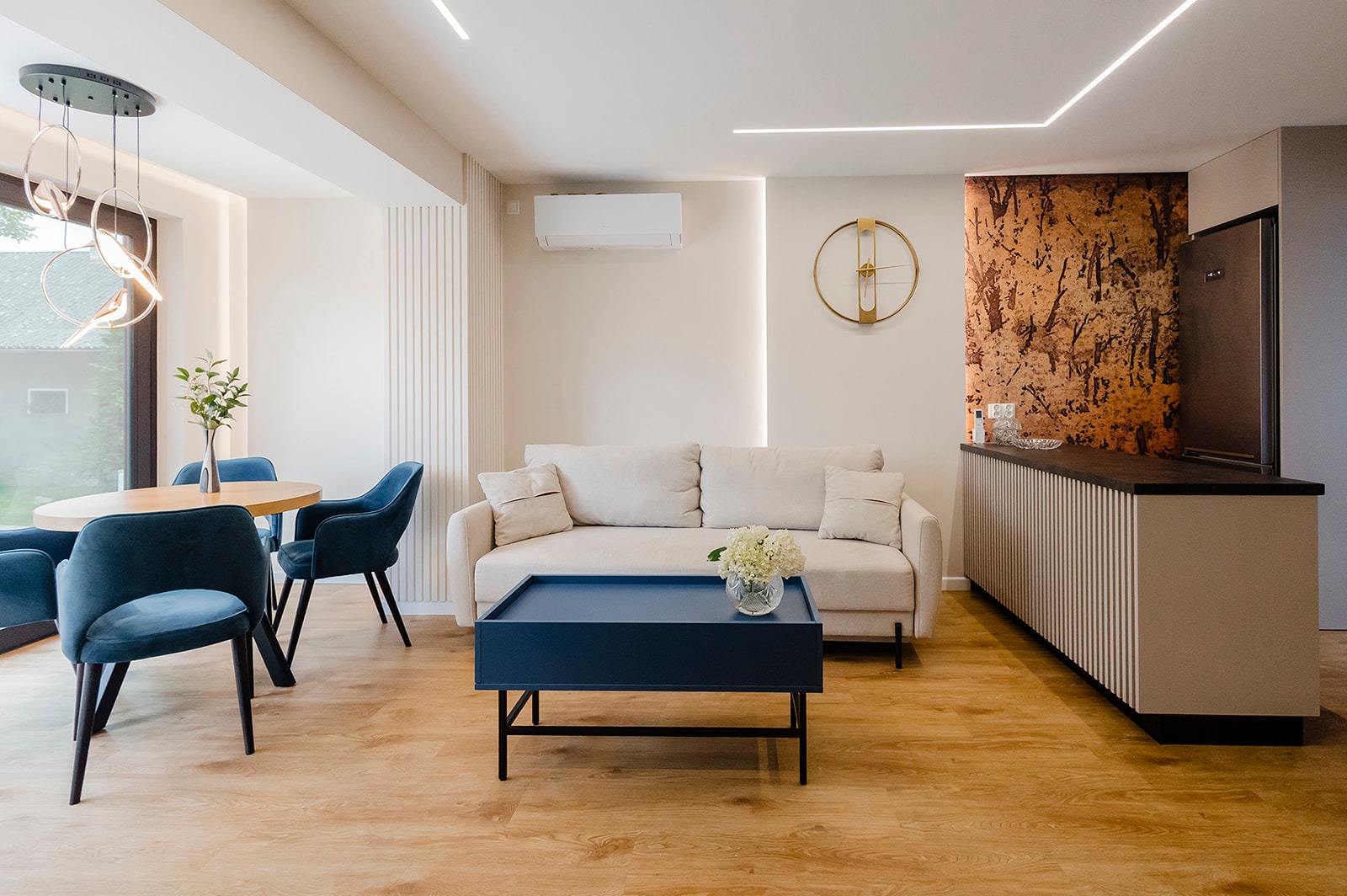 Jasne wnętrze z drewnianą podłogą, beżową sofą, okrągłym stołem i niebieskimi krzesłami. Na ścianie złoty zegar i dekoracyjny panel. Oświetlenie liniowe LED.