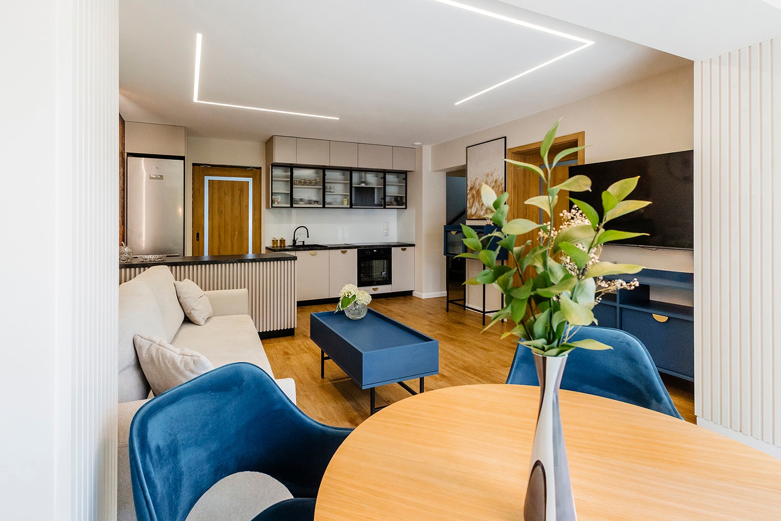 Nowoczesne wnętrze salonu z aneksem kuchennym w Jędrzejowie. Jasna sofa, niebieskie akcenty, drewniany stół i podłoga. Minimalistyczne oświetlenie sufitowe.