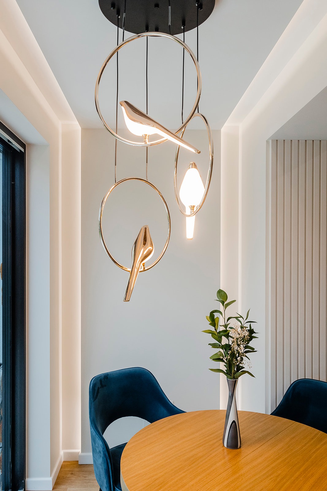 Nowoczesna lampa wisząca z motywem ptaków nad okrągłym stołem i welurowymi krzesłami w jadalni. Minimalistyczny design z akcentami złota i subtelnym oświetleniem.