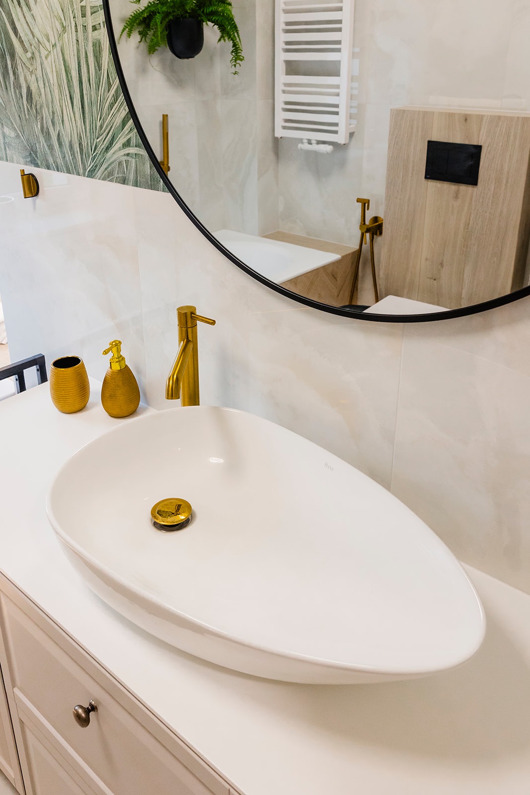 Biała, owalna umywalka nablatowa z baterią i akcesoriami w kolorze złotym. W tle okrągłe lustro z odbiciem fragmentu łazienki. Minimalistyczny design.