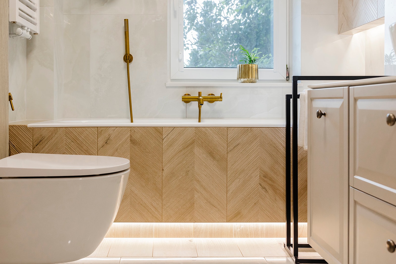 Jasna łazienka z drewnianą obudową wanny ułożoną w jodełkę, złotą armaturą i rośliną w złotej doniczce na parapecie. Widoczna toaleta i szafka łazienkowa.