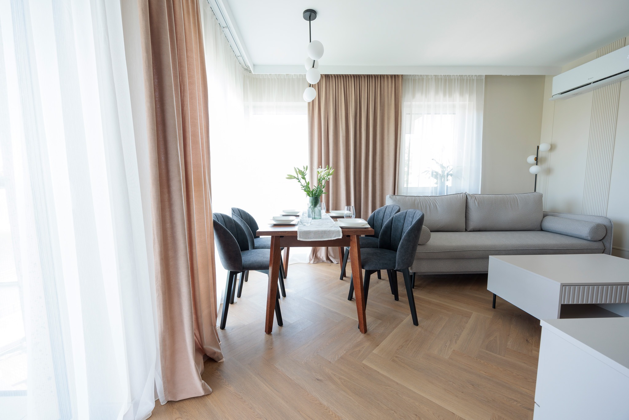 Jasne wnętrze: stół z szarymi krzesłami, sofa, parkiet w jodełkę, delikatne zasłony i nowoczesna lampa. Minimalistyczny design z akcentami beżu i szarości.