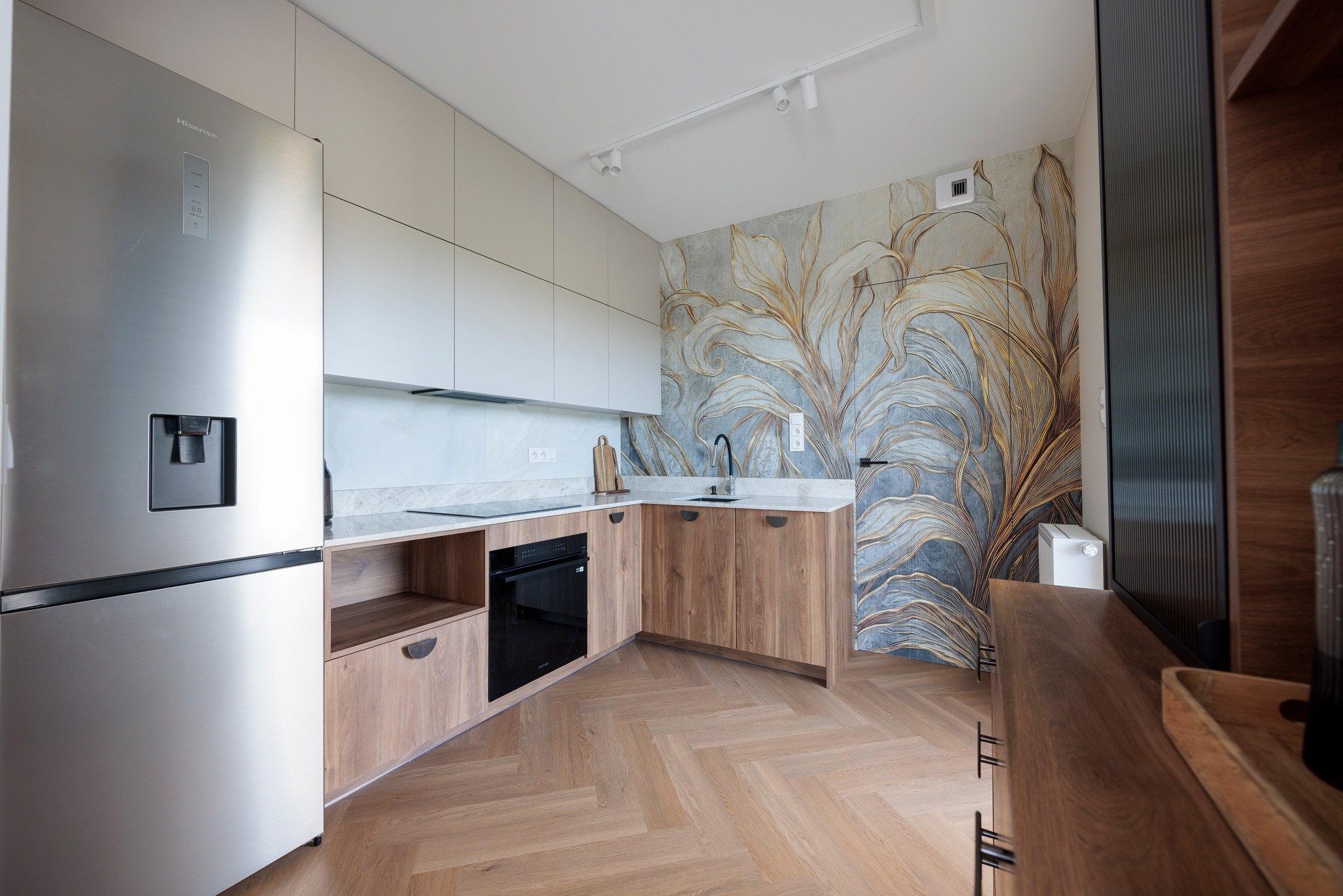 Nowoczesna kuchnia z drewnianymi szafkami, srebrną lodówką i dekoracyjną tapetą w odcieniach złota i szarości. Parkiet ułożony w jodełkę. Minimalistyczne oświetlenie.