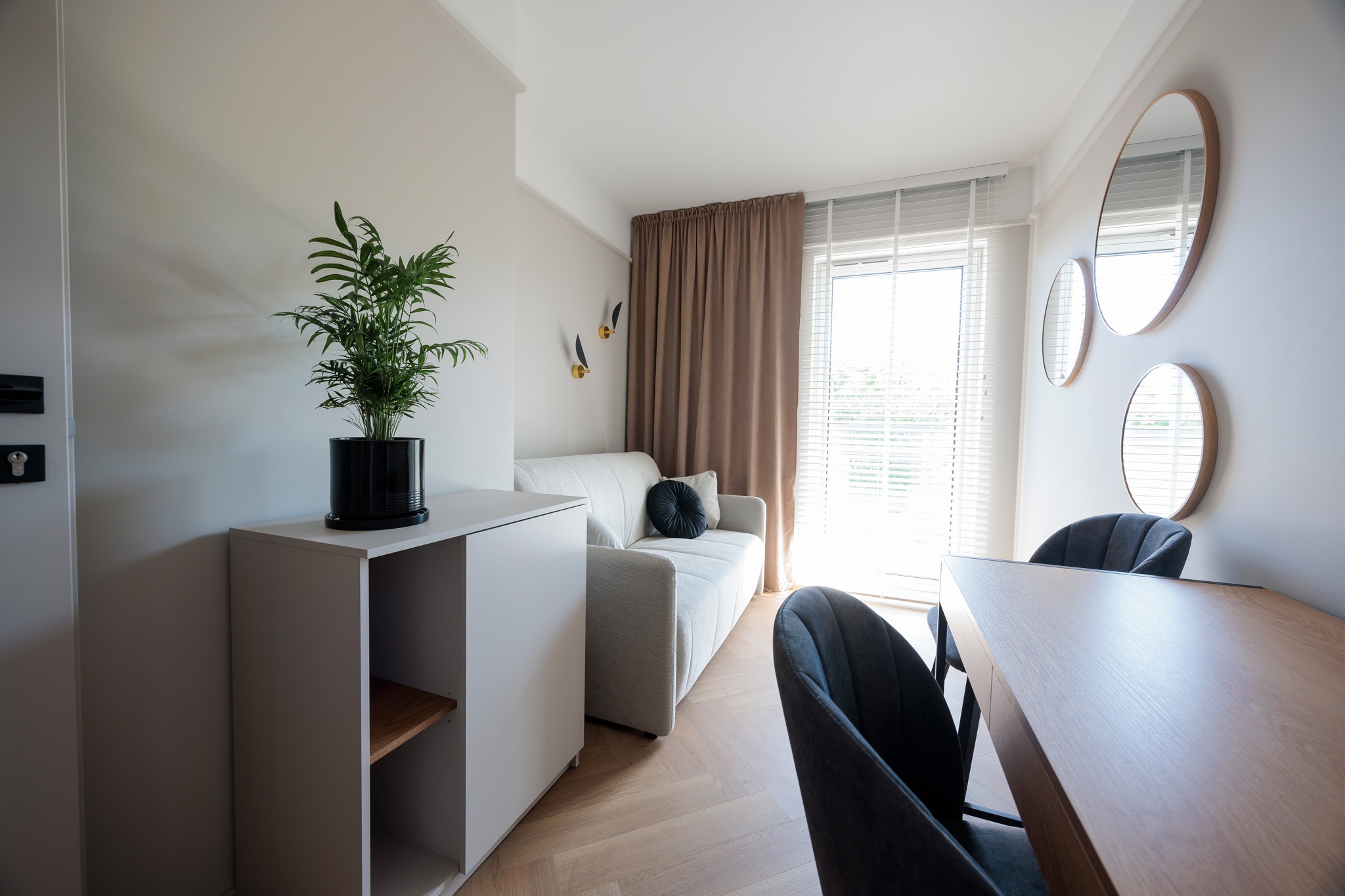 Jasne wnętrze w Jędrzejowie: sofa, stół z krzesłami, komoda z rośliną, okrągłe lustra. Minimalistyczny design, parkiet w jodełkę, naturalne światło.