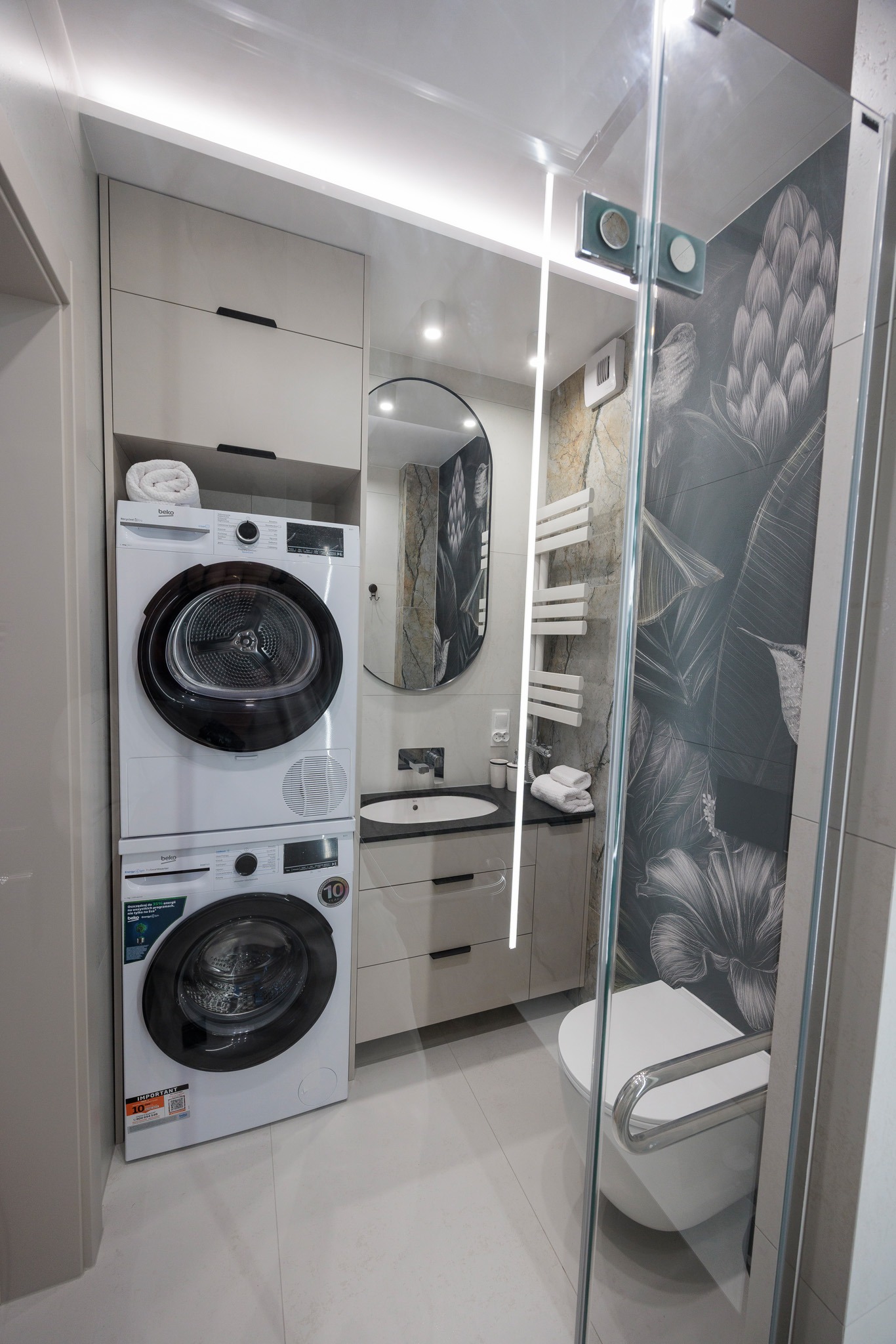 Nowoczesna łazienka z pralką i suszarką w zabudowie, owalne lustro, szklana kabina prysznicowa z motywem roślinnym na ścianie. Jasne płytki i meble.