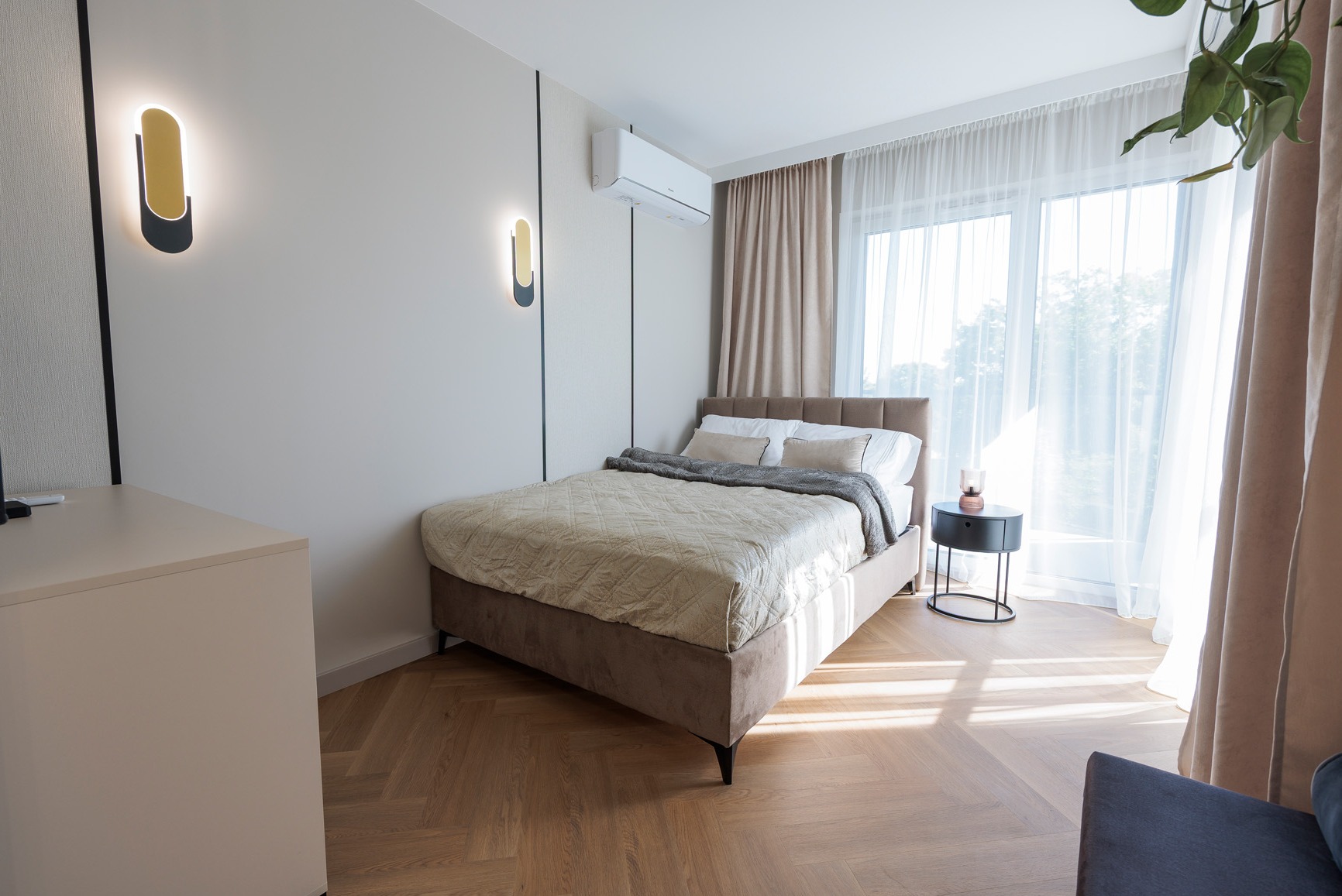 Nowoczesna sypialnia z łóżkiem w odcieniach beżu, drewnianą podłogą w jodełkę i minimalistycznymi lampami ściennymi. Jasne zasłony wpuszczają naturalne światło.