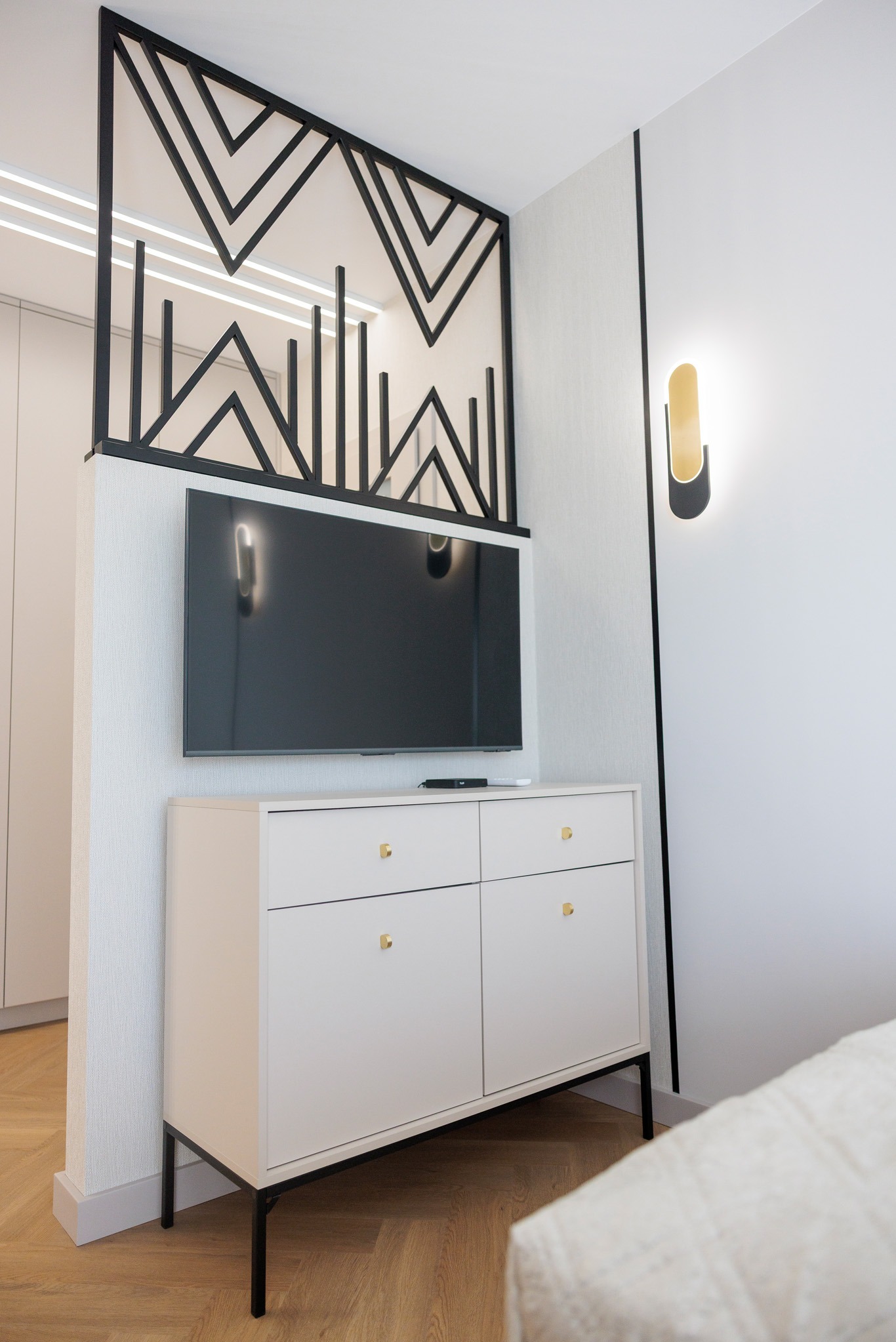 Nowoczesny pokój z białą komodą na czarnych nóżkach, telewizorem i dekoracyjną, czarną ścianką działową z geometrycznym wzorem. Minimalistyczna lampa ścienna.