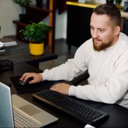 Krystian Saladra - Mężczyzna z brodą pracuje przy biurku z laptopem i klawiaturą. W tle roślina w żółtej doniczce i schody. Ujęcie z perspektywy lekko z góry.