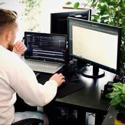 Krystian Saladra - Programista pracuje przy biurku z dwoma monitorami, na jednym z nich widoczny kod. Rośliny w tle dodają naturalnego charakteru przestrzeni pracy.