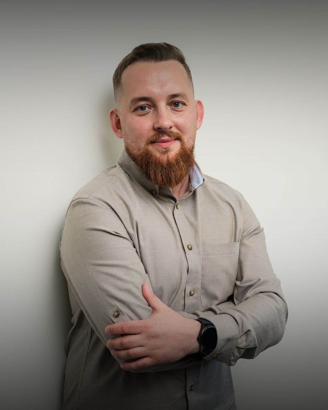 Portret brodatego mężczyzny w beżowej koszuli, z założonymi rękami i zegarkiem na nadgarstku, stojącego na tle jasnej ściany. Uśmiechnięty, z krótką fryzurą.