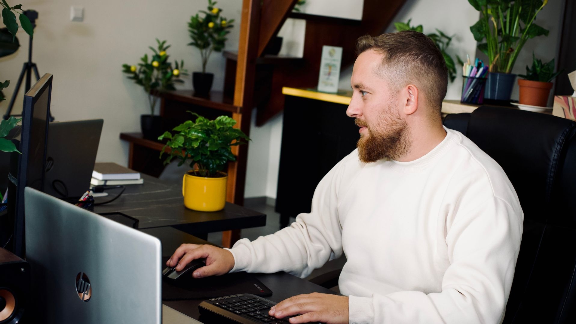 Mężczyzna z brodą pracuje przy biurku z dwoma laptopami, monitorem i klawiaturą. W tle schody i rośliny doniczkowe. Widok z boku, skupienie na pracy.