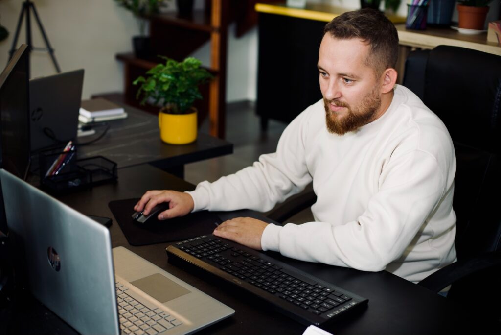 Mężczyzna z brodą pracuje przy biurku z laptopem i klawiaturą. W tle roślina w żółtej doniczce i schody. Ujęcie z perspektywy lekko z góry.