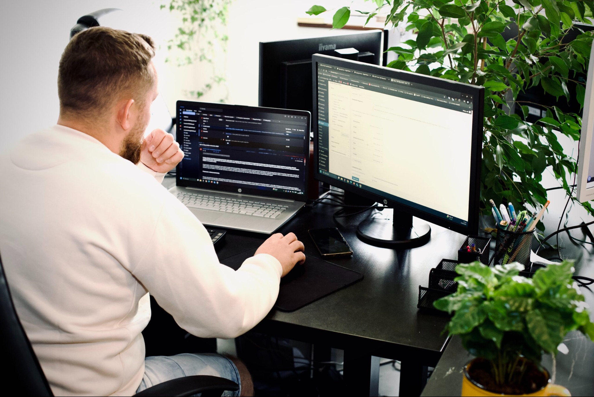 Programista pracuje przy biurku z dwoma monitorami, na jednym z nich widoczny kod. Rośliny w tle dodają naturalnego charakteru przestrzeni pracy.