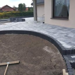 KW Grunt Tech Sp. z o.o. - Nowo ułożona szara kostka brukowa w Krerowie, tworząca zakrzywioną ścieżkę i taras przy domu, z ozdobnym obrzeżem z ciemnej palisady i dwoma stopniami, obok przygotowanego podłoża.