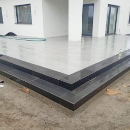 KW Grunt Tech Sp. z o.o. - Nowoczesny taras z lśniących, jasnoszarych płytek ceramicznych i dwoma ciemnymi, eleganckimi stopniami w kształcie litery L, prowadzącymi do surowego gruntu przy białym budynku.