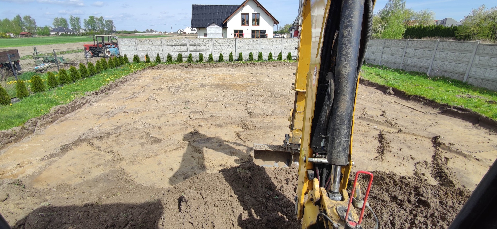 Żółta koparka w trakcie prac ziemnych na działce, widoczny wykop pod fundamenty, ogrodzenie z prefabrykatów betonowych i dom w tle. Dzień słoneczny.