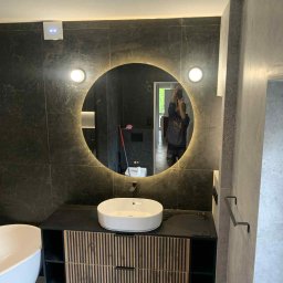 Wnętrza z pasją - Nowoczesna łazienka z okrągłym lustrem LED, umywalką nablatową i szafką z lamelami. W lustrze odbija się osoba robiąca zdjęcie. Ciemna glazura na ścianach.