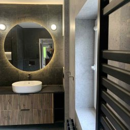 Wnętrza z pasją - Nowoczesna łazienka z okrągłym lustrem podświetlanym LED, umywalką nablatową i grzejnikiem drabinkowym. Szare płytki i drewniane akcenty szafki pod umywalką.