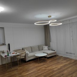 Wnętrza z pasją - Jasne wnętrze salonu z drewnianą podłogą, beżową sofą i okrągłymi lampami sufitowymi. Ściana z dekoracyjnymi panelami i subtelne zasłony w tle.