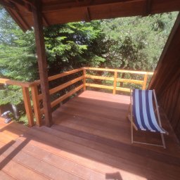 Wnętrza z pasją - Drewniany taras z barierką i krzesłem plażowym w paski, otoczony zielenią. Widoczne narzędzia sugerują niedawne prace wykończeniowe.