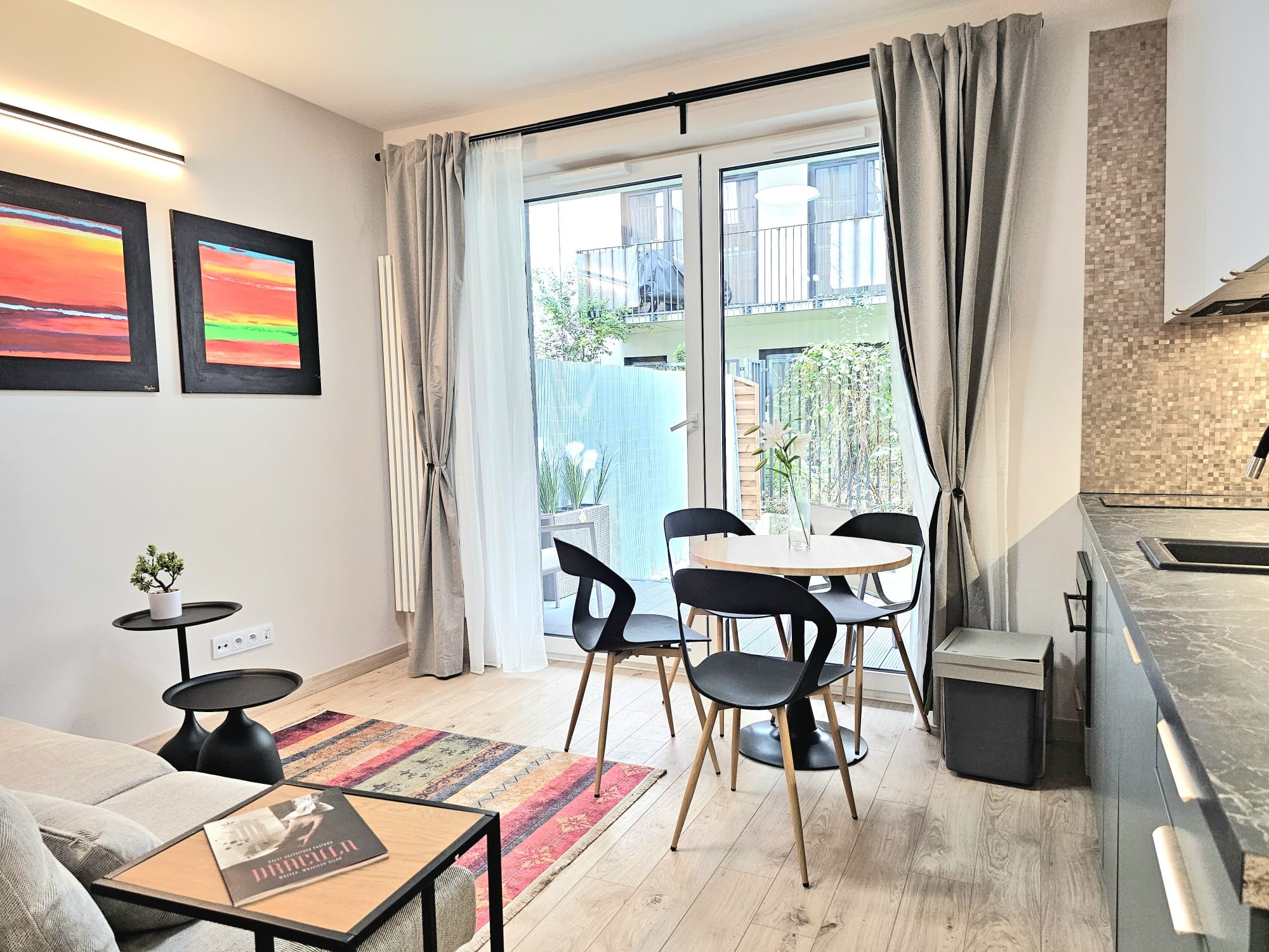 Jasne wnętrze mieszkania z aneksem kuchennym, okrągłym stołem i wyjściem na balkon. Na ścianie wiszą obrazy w czarnych ramach. Parkiet na podłodze.