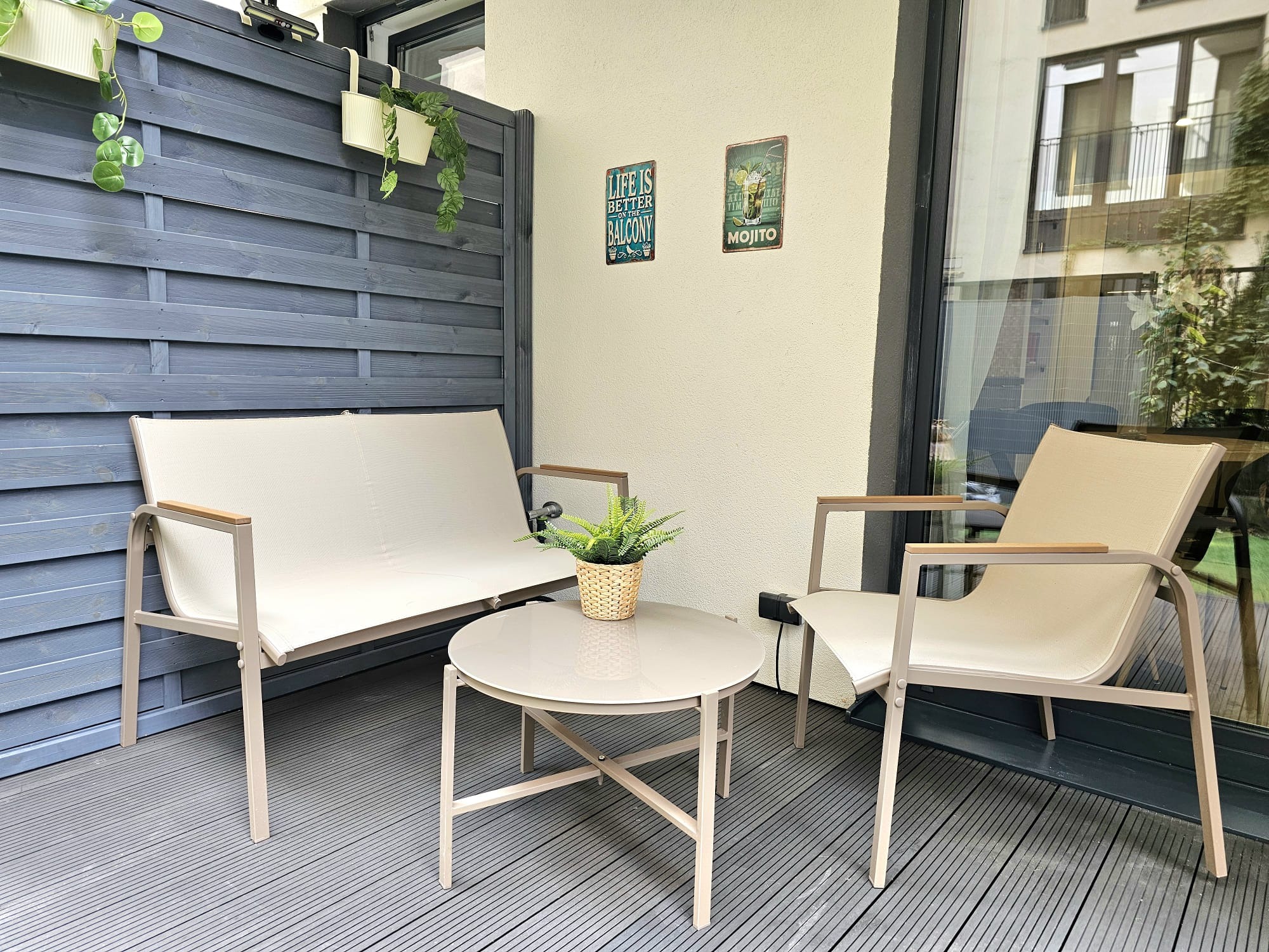 Przytulny balkon z szarą drewnianą ścianą, beżowymi meblami ogrodowymi i dekoracjami ściennymi. Rośliny doniczkowe dodają świeżości.