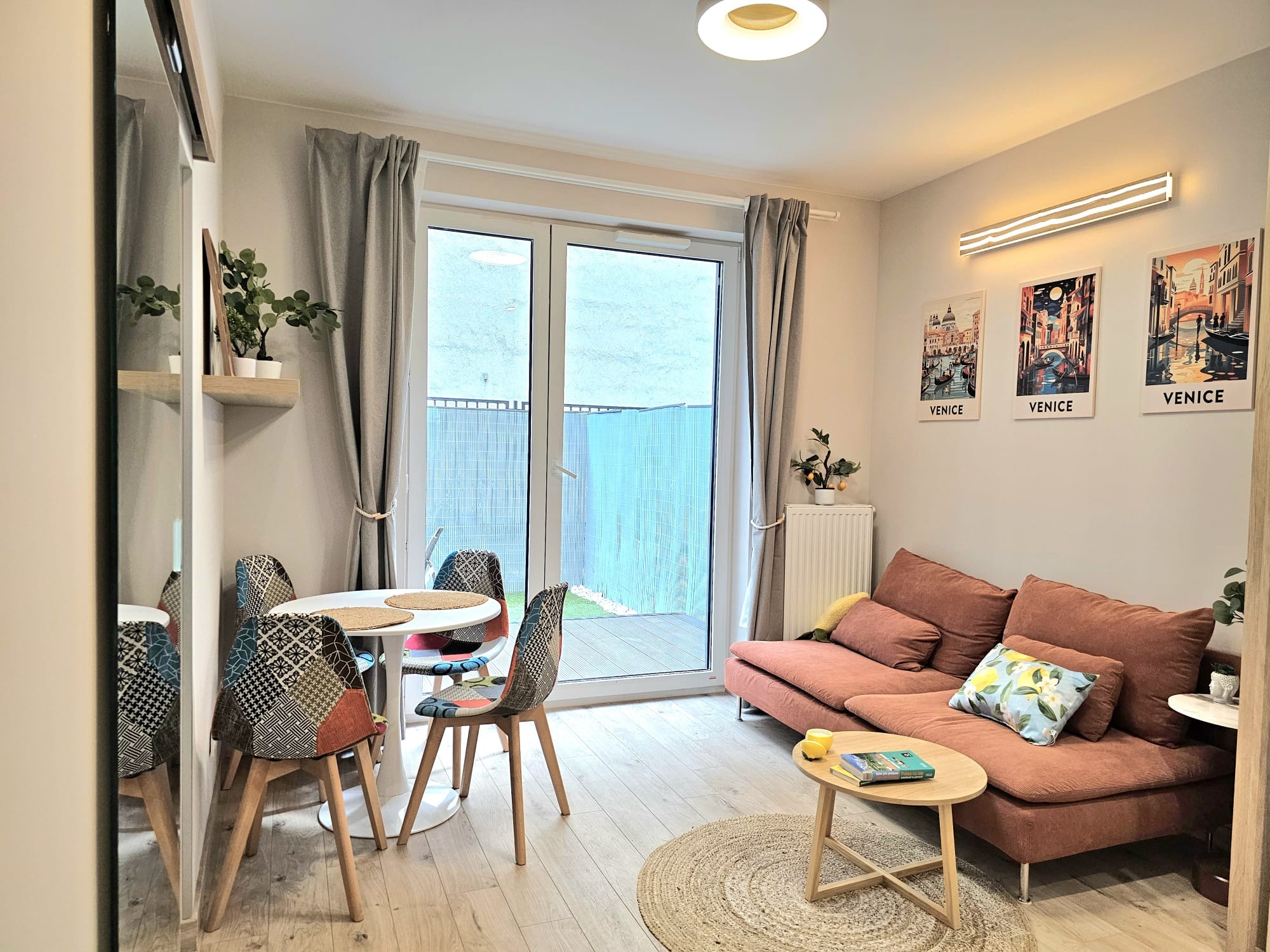 Jasne wnętrze salonu z okrągłym stołem i patchworkowymi krzesłami, kanapą w odcieniach różu, drewnianą podłogą i plakatami z motywem Wenecji na ścianie.