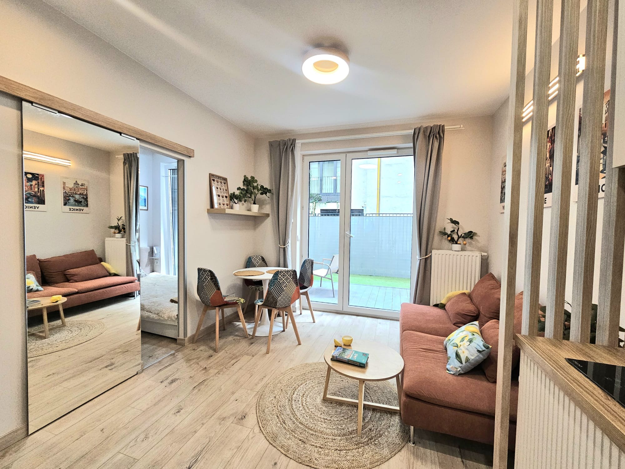 Jasne wnętrze salonu z drewnianą podłogą, rozsuwanymi drzwiami z lustrem i wyjściem na balkon. Widoczny stół z krzesłami, sofa i dekoracyjna ścianka działowa.