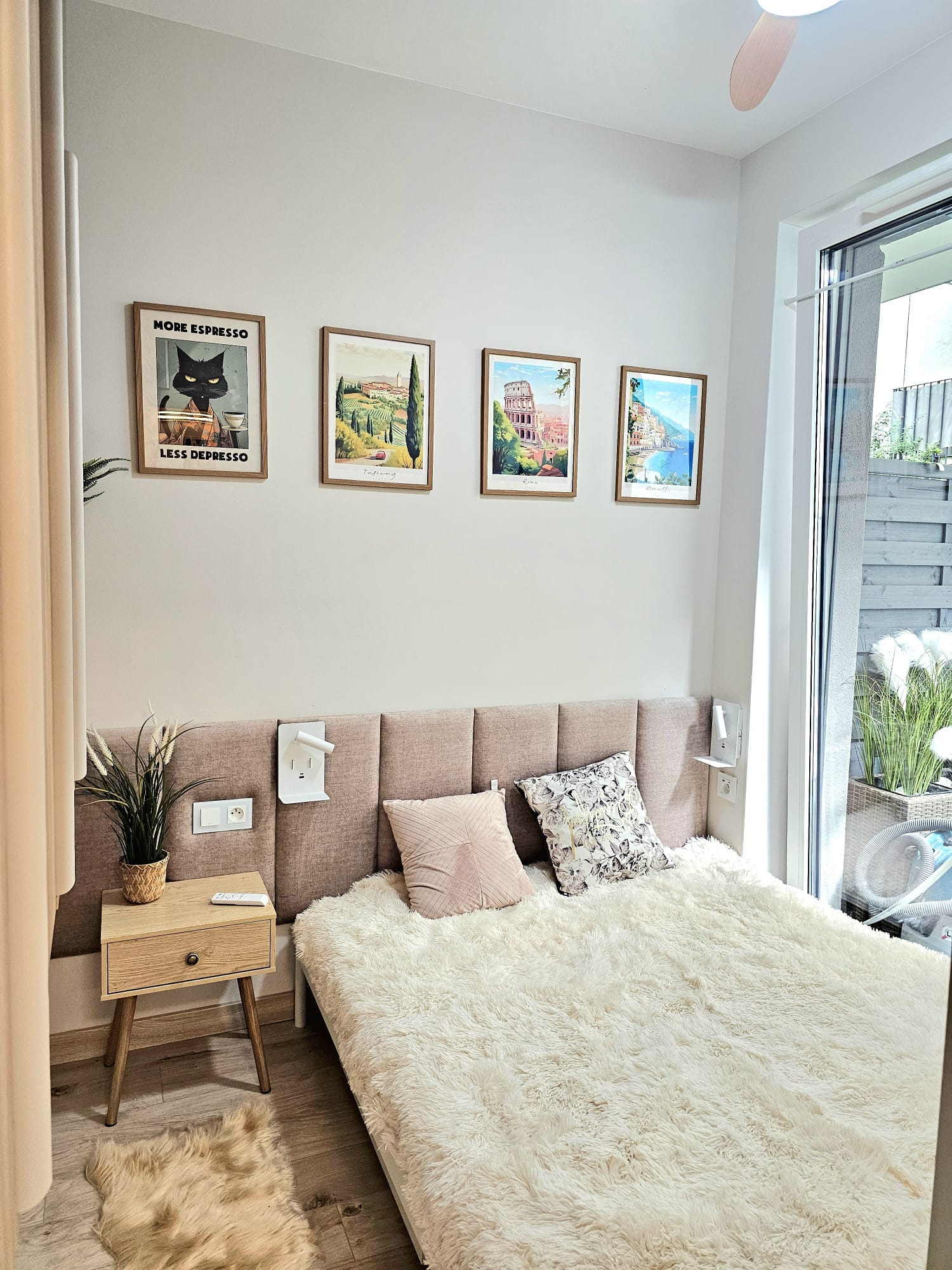 Jasna sypialnia z łóżkiem, stolikiem nocnym i dekoracjami ściennymi w postaci obrazków z motywami włoskimi. Widok na balkon z roślinami.